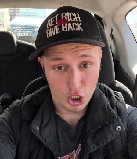 A man sitting in a car making an exaggerated funny expression, wearing a black vest over a grey t-shirt and a black cap with the text "GET RICH GIVE BACK" and a small red animal icon.